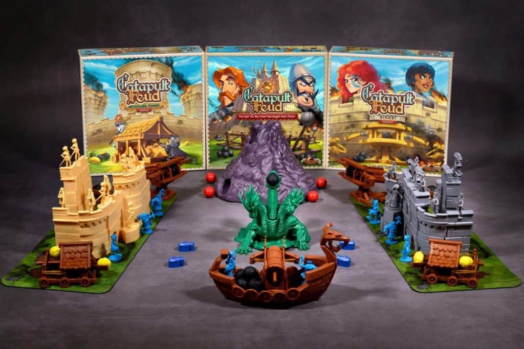 A tabletop game setup showcases the thrilling "Catapult Feud" board game with two opposing castle structures, each armed with small catapults and pieces. Central to the scene is a majestic green dragon on a boat. Three vibrant game boxes flank the background, reminiscent of an animated series.