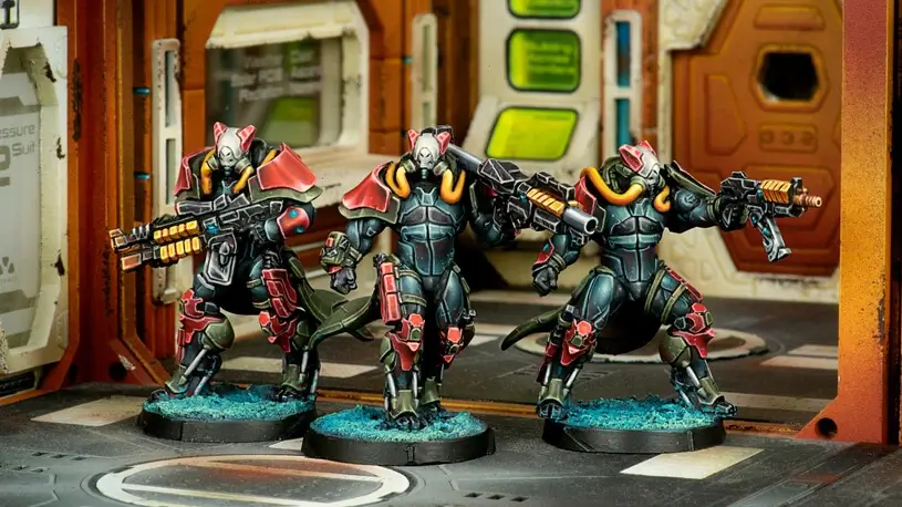 In June's new releases, three futuristic, armored figures with animal-like helmets and large weapons dominate the sci-fi setting. Their predominantly black outfits feature striking red and orange accents, all while they stand poised on circular bases in an aura of Infinity.